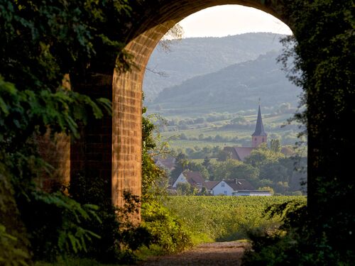 Blick durch einen Bogen auf ein Dorf mit Kirche, umgeben von Weinbergen und sanften Hügeln. | © Name