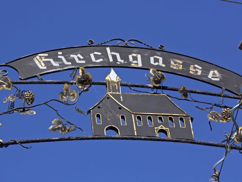 Das dekorative Schild "Kirchgasse" zeigt eine Kirche vor strahlend blauem Himmel. | © Name