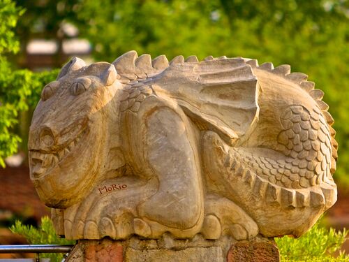 Eine kunstvolle Holzskulptur eines Ungeheuers liegt in einem grünen Forst. | © Morio