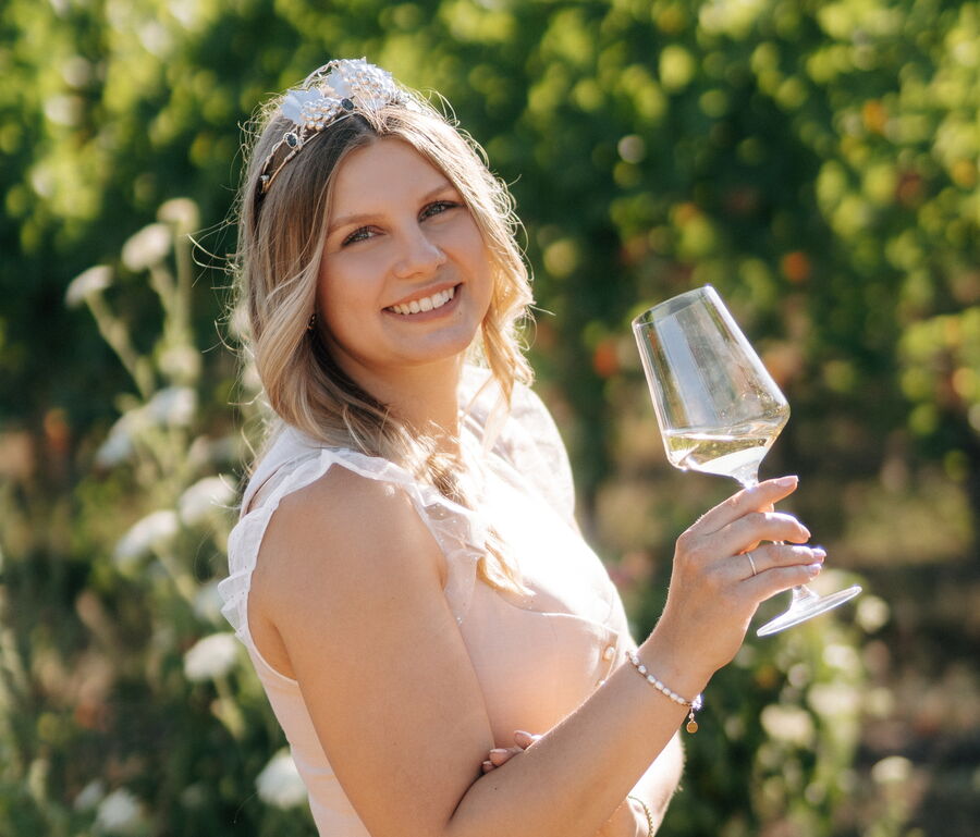 Unsere Verbandsgemeinde Prinzessin Deidesheim hält ein Weinglas vor einer grünen Weinbergkulisse in der Sonne.