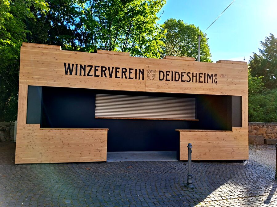 Holzstand des Winzervereins Deidesheim mit großem Schriftzug, umgeben von Bäumen und gepflastertem Boden. | © T-I