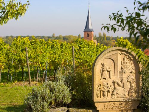 Weinberge mit einer Steinskulptur im Vordergrund und einem Kirchturm im Hintergrund in der Forst Weinlage Kirchenstück.