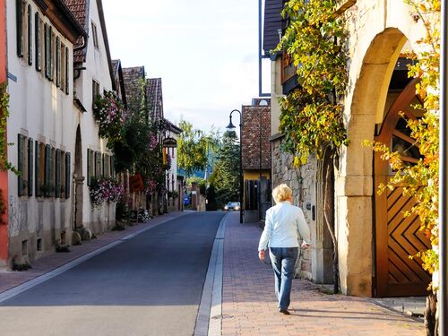 Eine Frau spaziert entlang der malerischen Straßen von Ruppertsberg, umgeben von blühenden Pflanzen und historischen Gebäuden.