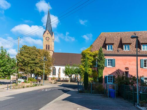 Die Kirche in Ruppertsberg erhebt sich majestätisch über die ruhige Straße an einem sonnigen Tag. | © Name