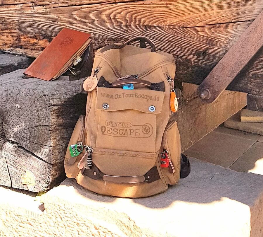 Ein brauner Rucksack steht neben einem Notizbuch auf einem hölzernen Untergrund. | © OnTourEscape