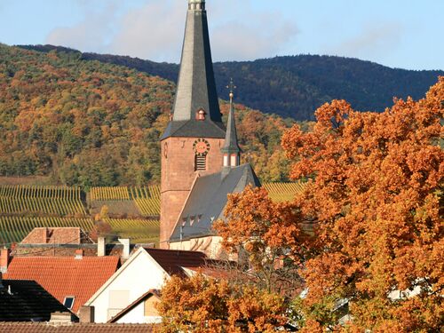 eine gotische Kirchturmspitze umgeben von bunt belaubten Weinbergen und Pfälzerwald