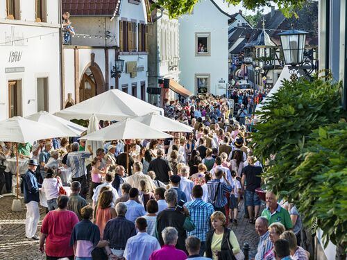 Menschenmengen genießen die Deidesheimer Weinkerwe unter Sonnenschirmen in einer lebhaften Straße. | © Name