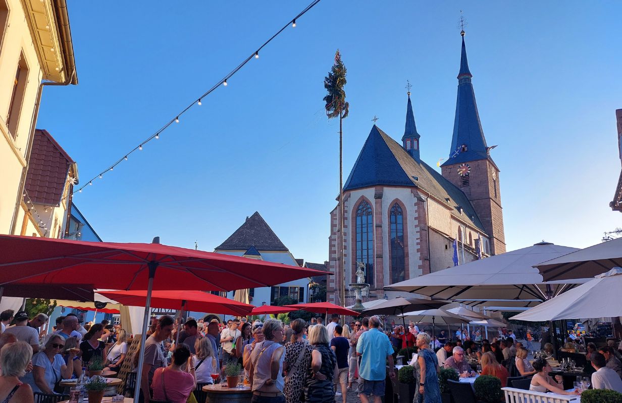 Belebte Deidesheimer Weinkerwe mit Menschen, roten Schirmen und der beeindruckenden Kirche im Hintergrund.
