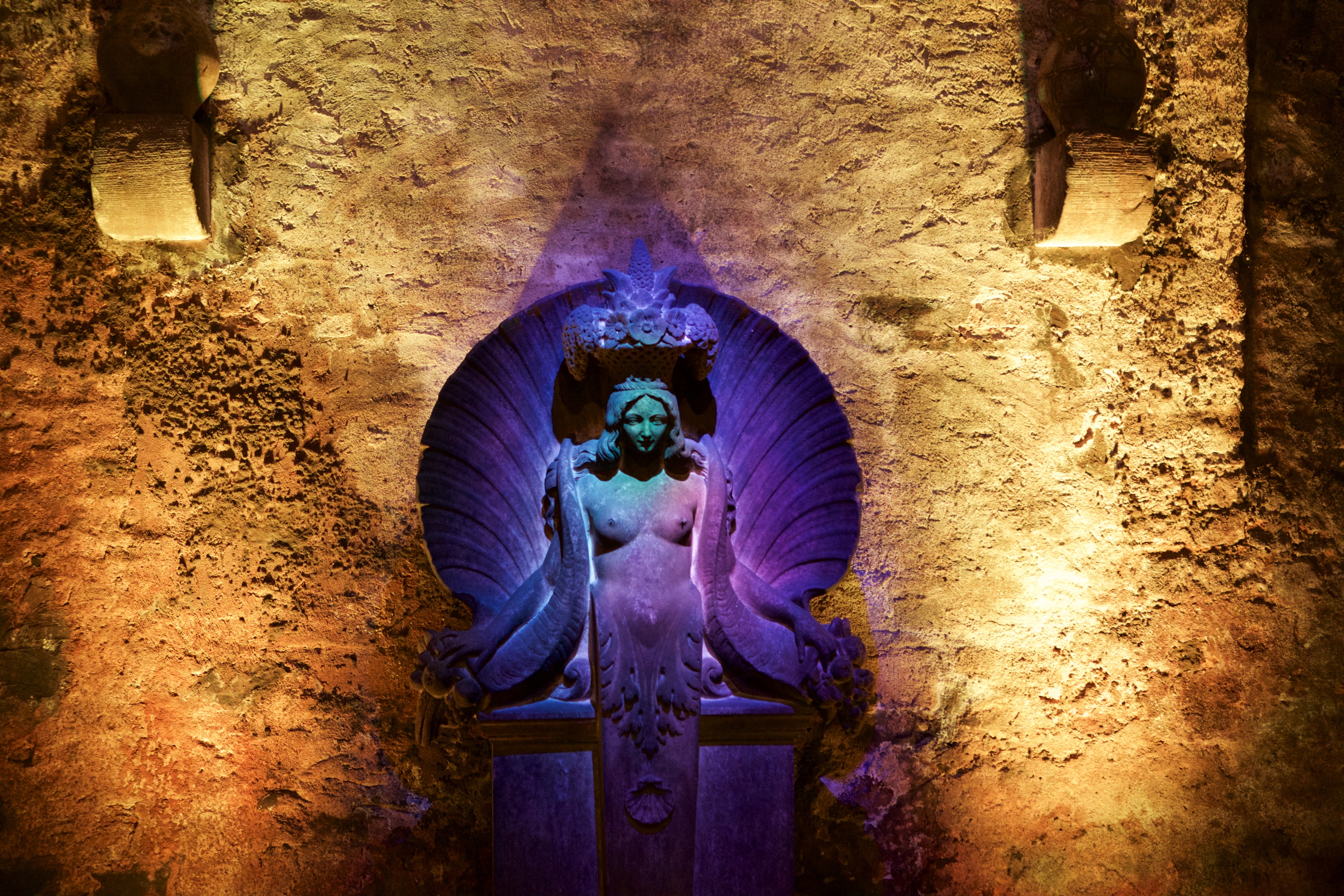 Eine kunstvolle Statue, beleuchtet vor einer strukturierten Wand, zeigt eine feminine Form in einer Muschel.