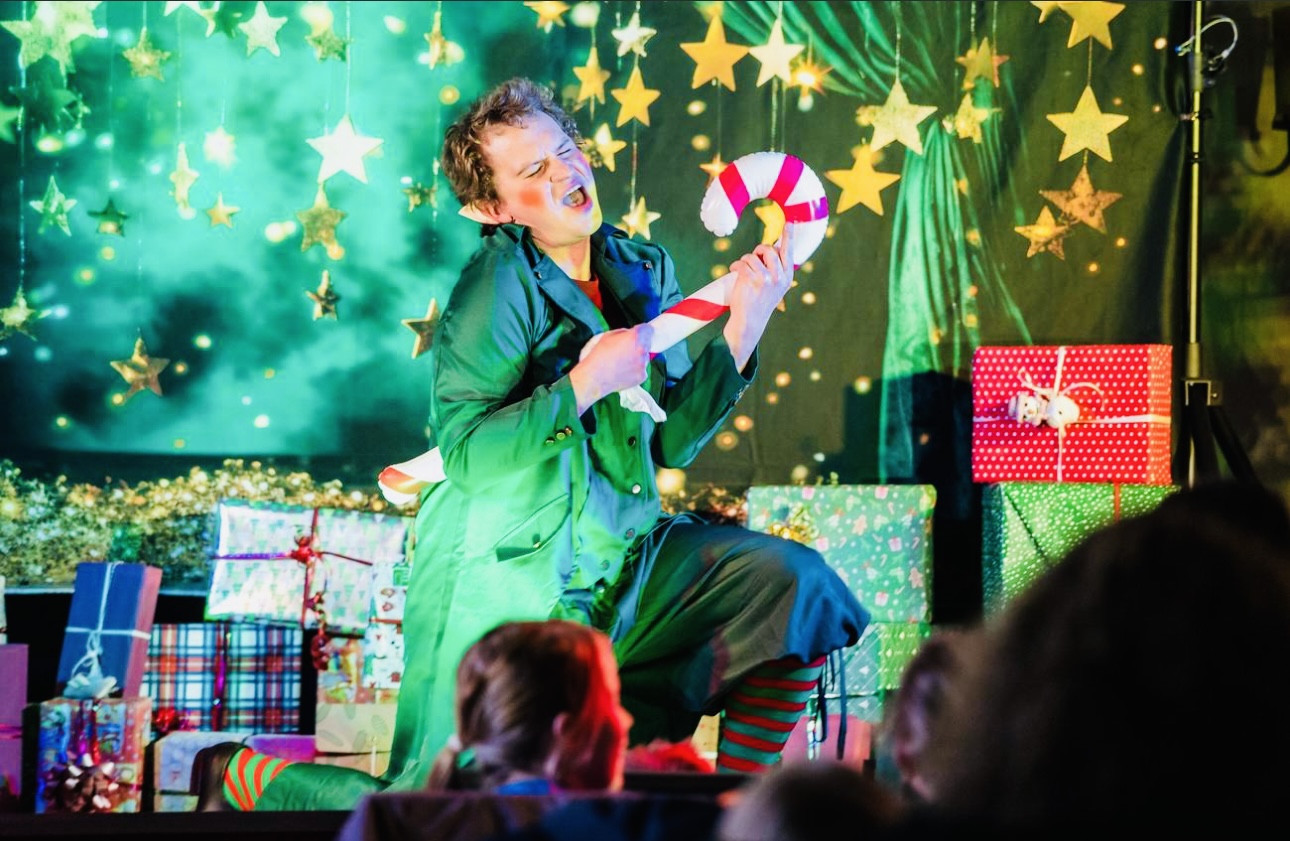 Ein Elf singt fröhlich mit einem Zuckerstab vor Weihnachtsgeschenken und leuchtenden Sternen.