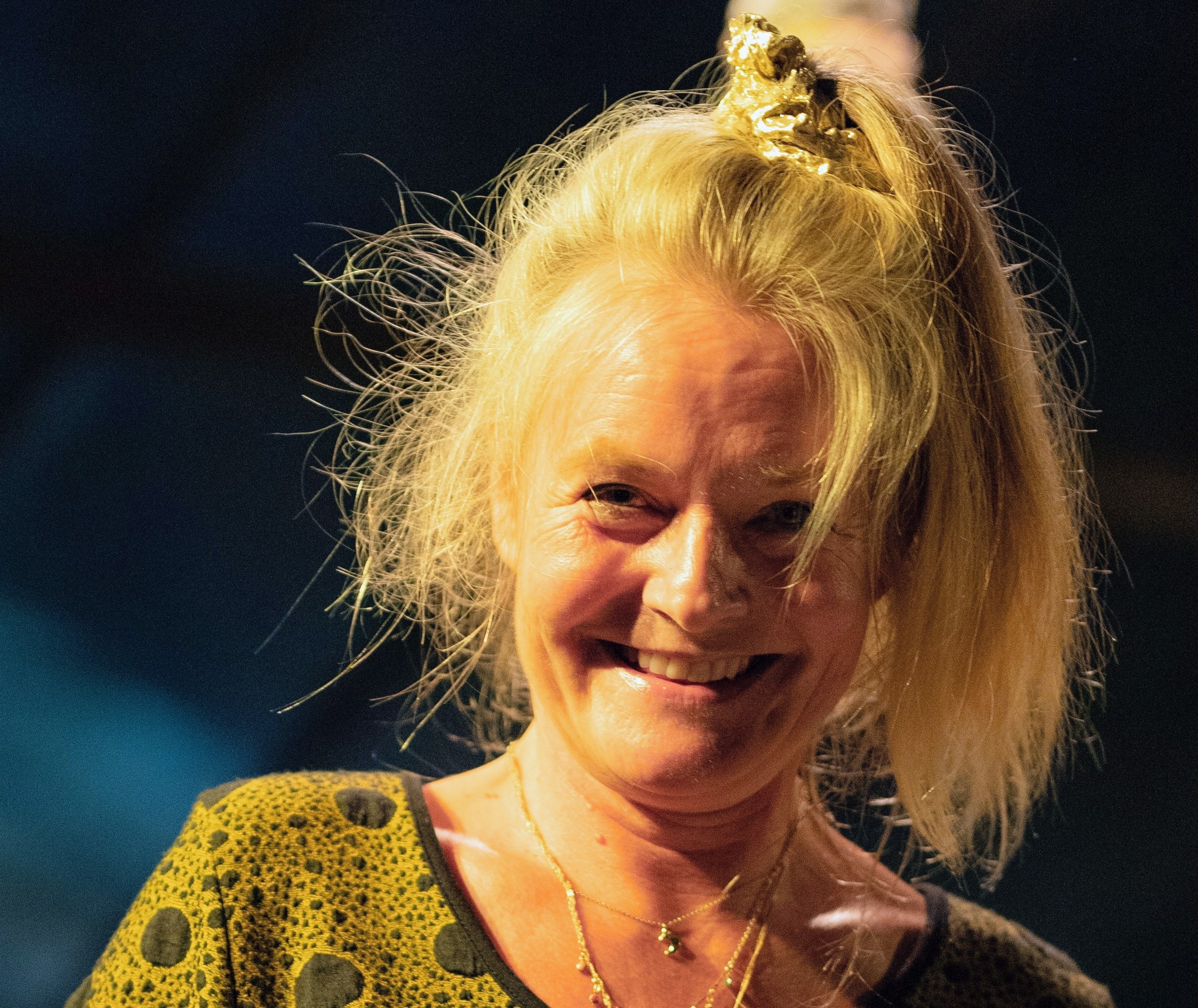Eine fröhliche Frau mit unordentlichem Haar und goldener Haarschmuck lächelt strahlend in die Kamera.